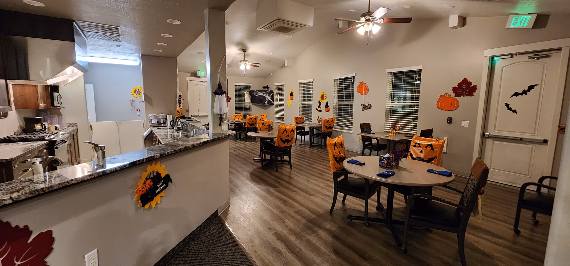 Interior view of a senior living facility dining room decorated for Halloween with pumpkin-themed chair covers, wall decorations including bats, pumpkins, and leaves. The room features round tables with chairs, a kitchen area with granite countertops, and ceiling fans with lights.