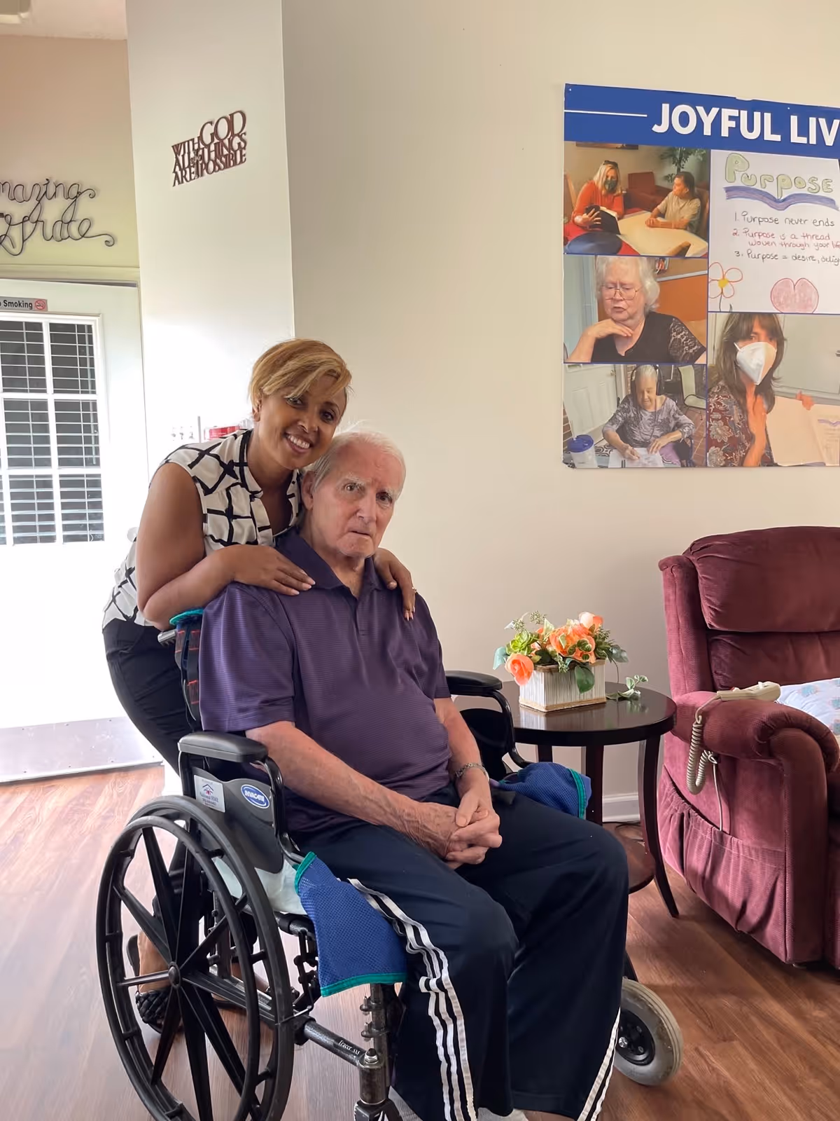 A woman stands behind an elderly man seated in a wheelchair inside a living area. The woman is smiling and has her arms gently resting on the man's shoulders. The room has wooden flooring, a small table with a flower arrangement, and a maroon recliner chair. On the wall behind them is a bulletin board with photos and the heading 'JOYFUL LIV'.