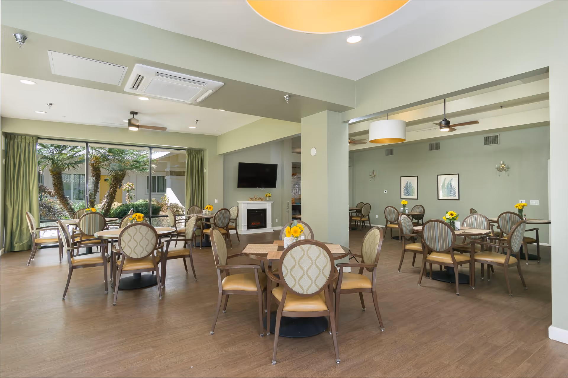 Sunlit communal dining room with round tables and chairs, large windows overlooking landscaping, a wall-mounted TV and a fireplace.