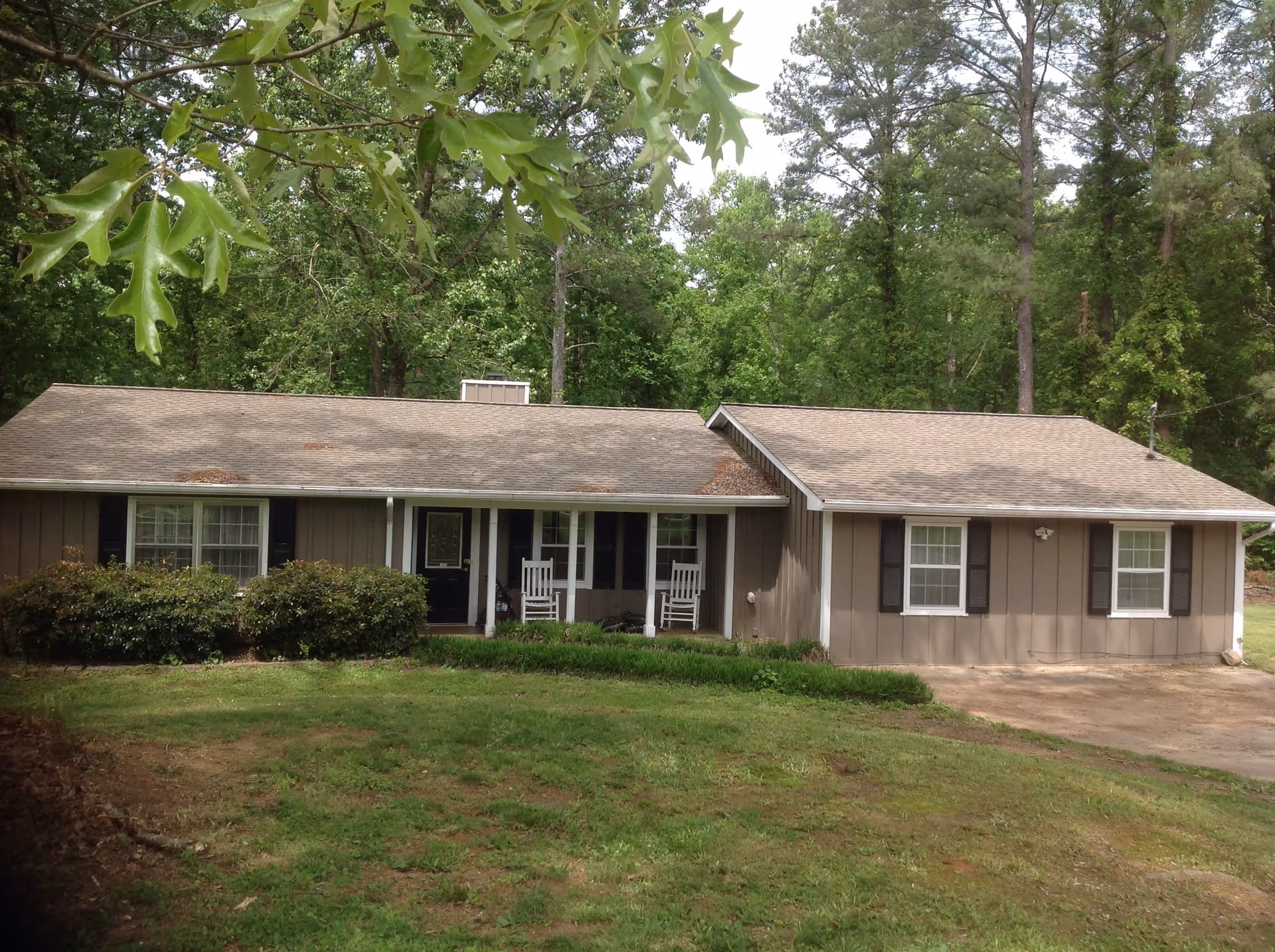 Single-story residential building with a brown exterior, black shutters, and a front porch featuring two white rocking chairs. The house is surrounded by green grass, bushes, and tall trees in the background.