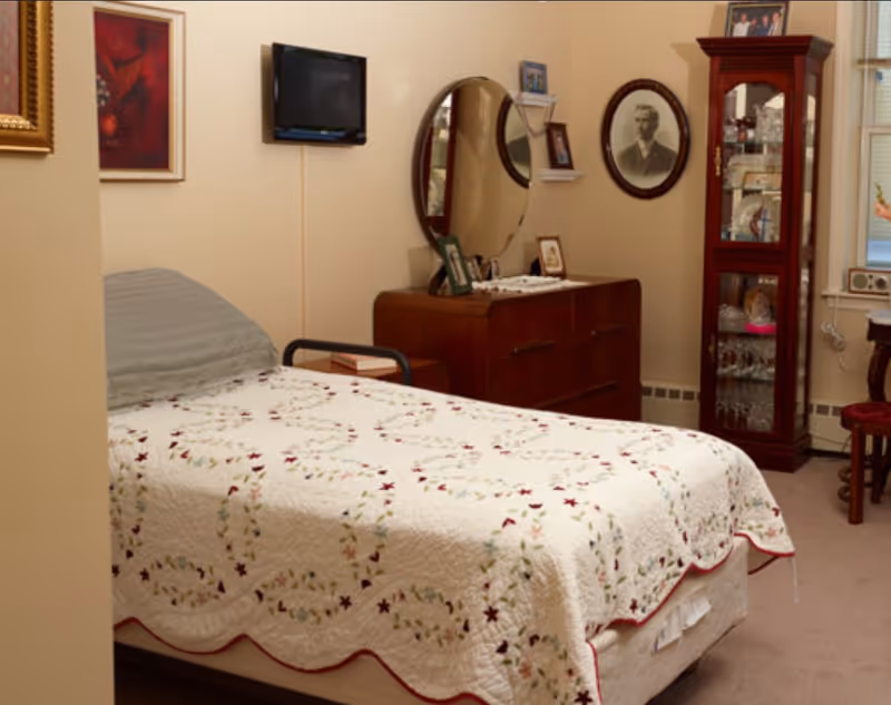 A bedroom with a single bed covered with a white quilt featuring a floral pattern. There is a wooden dresser with a large round mirror and framed photos on top. A tall glass-fronted cabinet filled with glassware stands next to a window with blinds. A small wall-mounted TV is above the bed, and there are framed pictures on the walls.