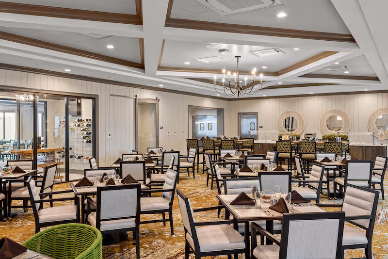 A spacious dining room with multiple tables and chairs arranged neatly. Each table is set with glasses and folded brown napkins. The room features a coffered ceiling with recessed lighting and a chandelier. There is a bar area with high chairs and round mirrors on the wall behind it. The walls are light-colored with wood paneling, and there is a glass door leading to another room.