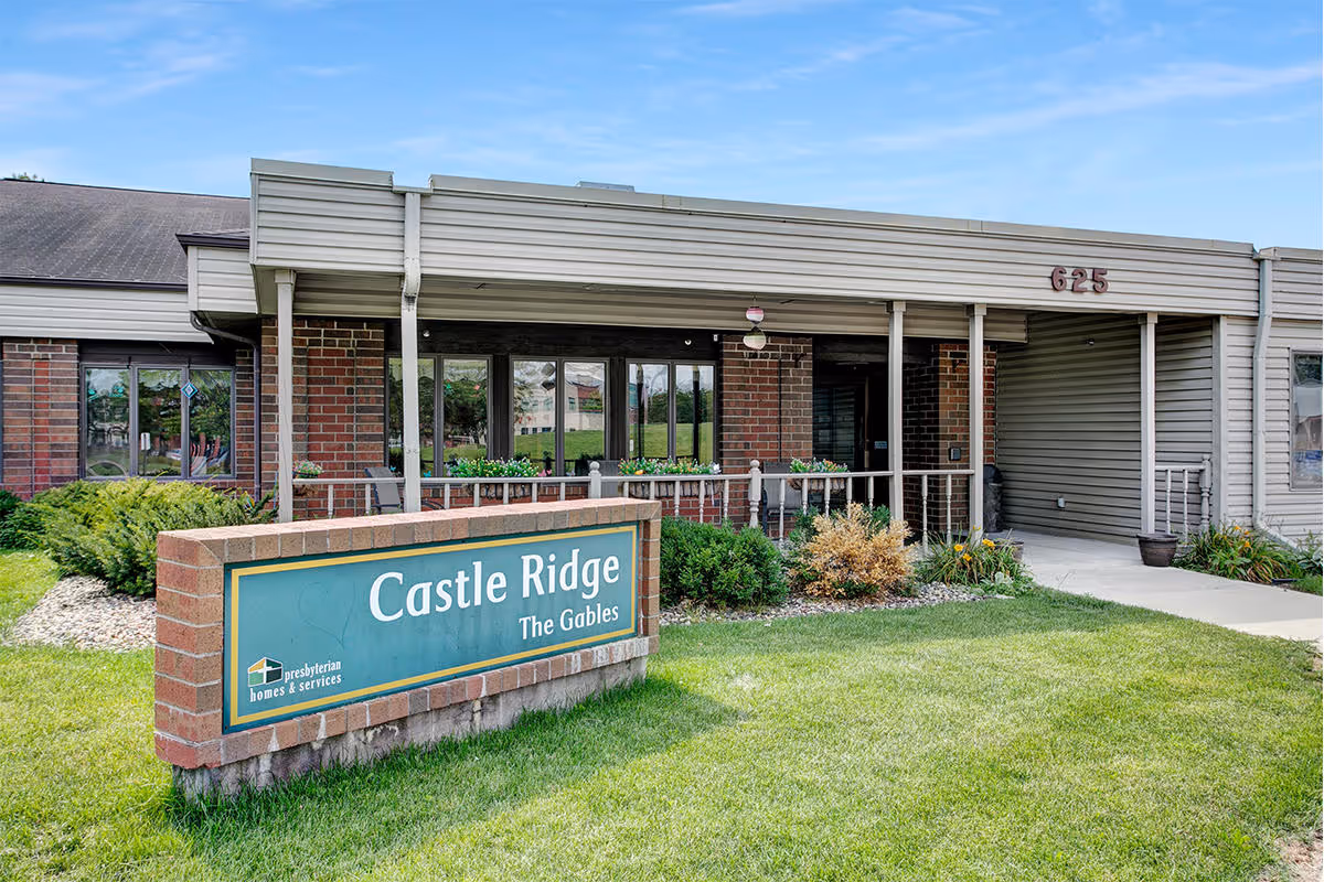 Exterior view of Castle Ridge senior living facility showing a brick and siding building with a covered entrance and a green sign in front that reads 'Castle Ridge The Gables' with landscaping and a clear blue sky.
