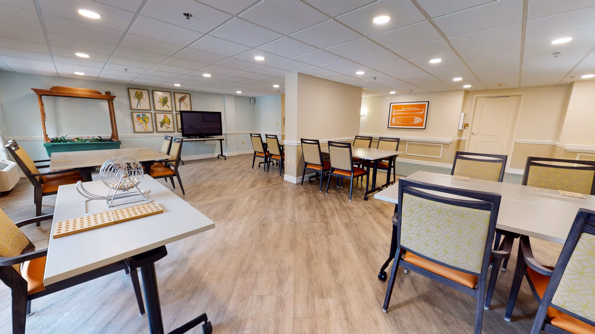 A spacious activity room with several tables and chairs arranged for group activities. One table has a bingo cage and bingo cards, indicating the room is used for games or social gatherings. The room has wood flooring, light-colored walls, framed artwork, and a flat-screen TV mounted on a stand. The ceiling has recessed lighting, and there is a large mirror on one wall.