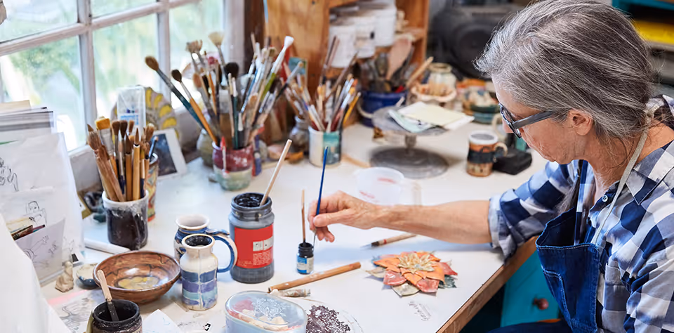 An elderly woman with gray hair and glasses is painting a decorative piece at a cluttered art workspace filled with paintbrushes, jars, and art supplies near a window.