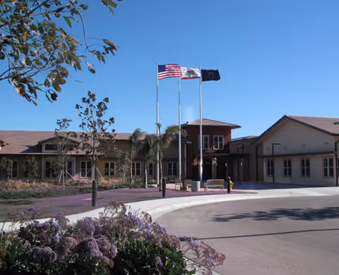 Front exterior of a single-story facility with three flagpoles and landscaped flowers under a clear blue sky.