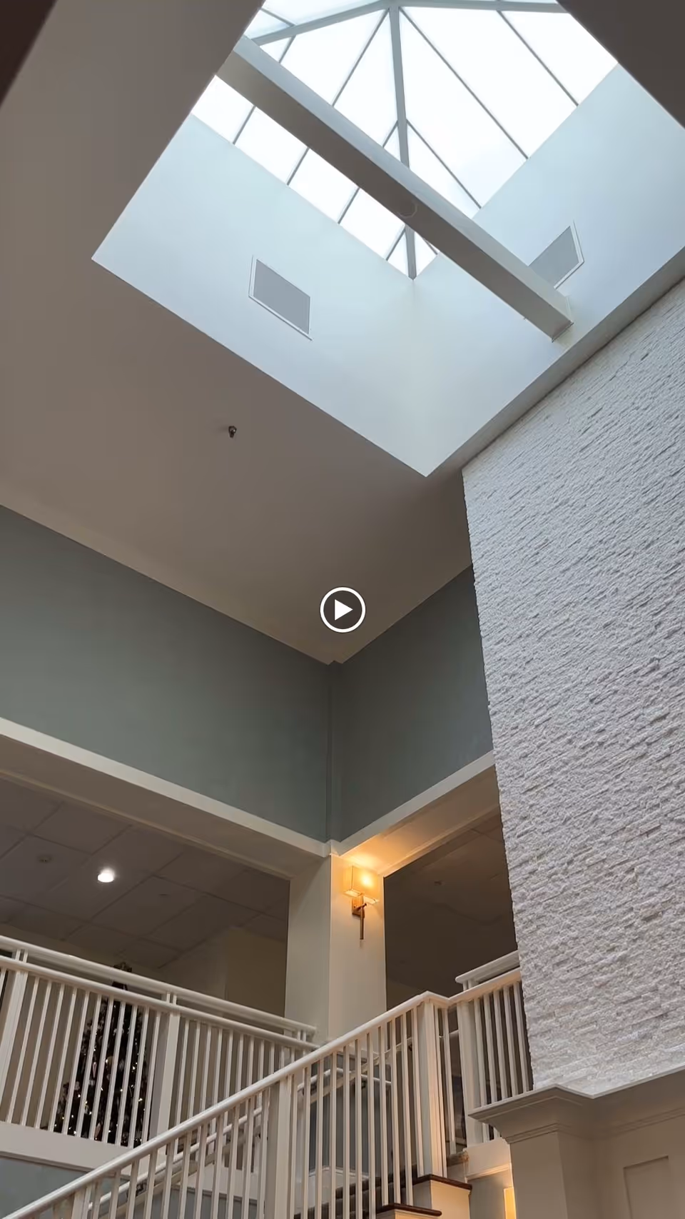 Interior view of a senior living facility showing a staircase with white railings leading up to a second floor. The walls are painted in muted tones with a textured white accent wall. A large skylight with a geometric frame allows natural light to illuminate the space. A wall-mounted light fixture is visible near the top of the stairs.