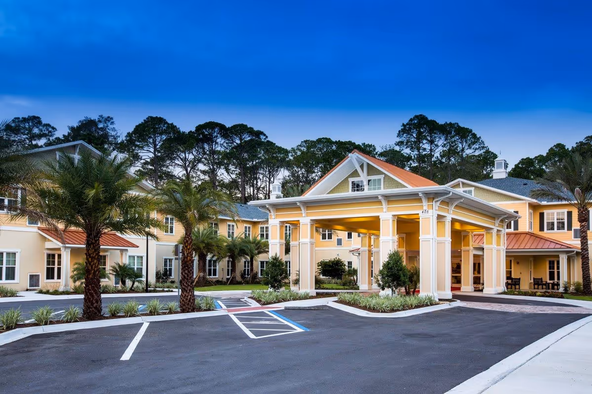 Exterior view of a senior living facility named Starling at Ponte Vedra, featuring a large covered entrance with white columns, yellow and beige building walls, palm trees, and a clear blue sky.