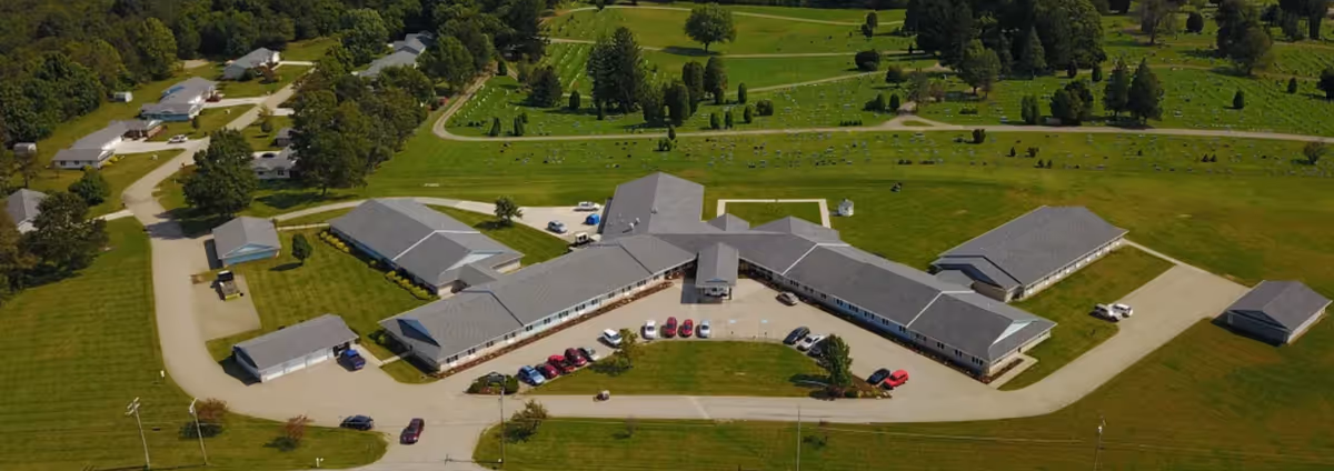 Aerial view of Woodcrest Senior Living Community showing a large, single-story building complex with multiple wings surrounded by green lawns and trees. Several cars are parked in front of the building, and there are paved roads and pathways around the facility. The surrounding area includes open grassy fields and a cemetery in the background.