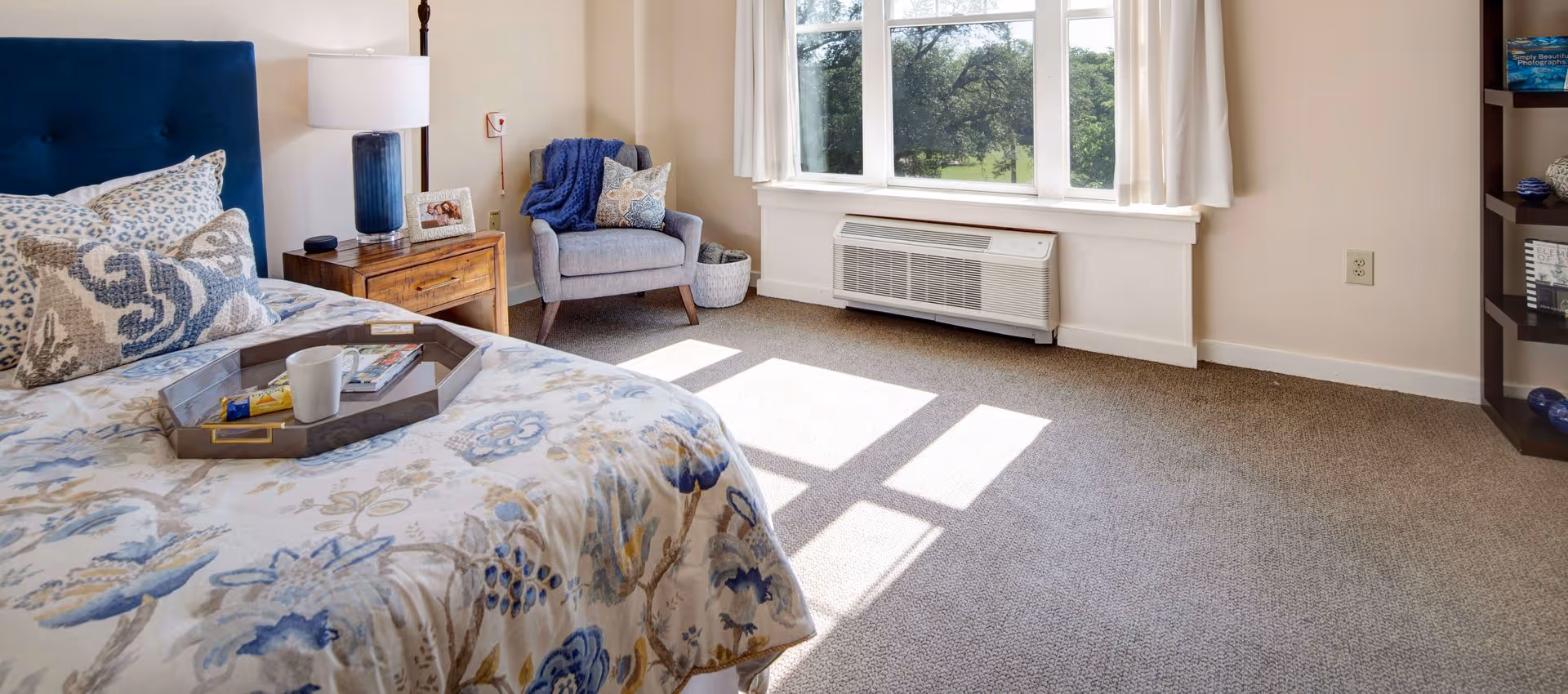 A bright bedroom with a bed featuring a blue upholstered headboard and floral bedding in shades of blue and beige. A wooden nightstand with a blue lamp and a framed photo sits next to the bed. There is a gray armchair with a blue throw blanket and a decorative pillow near a large window with white curtains, allowing natural light to fill the room. A basket is placed beside the chair, and a bookshelf with books and decorative items is visible on the right side.