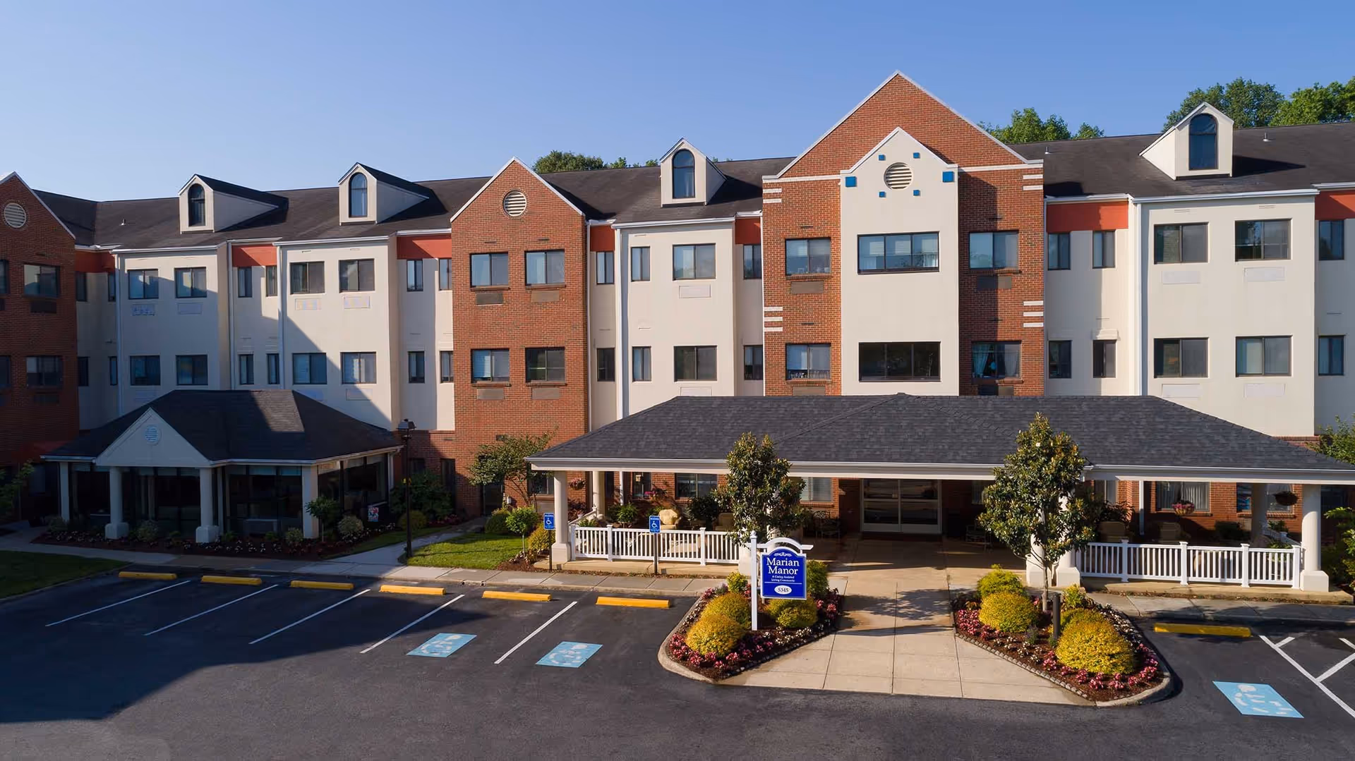 Front exterior view of Marian Manor Retirement Community building with a covered entrance, landscaped garden beds, and a parking lot with handicap spaces.