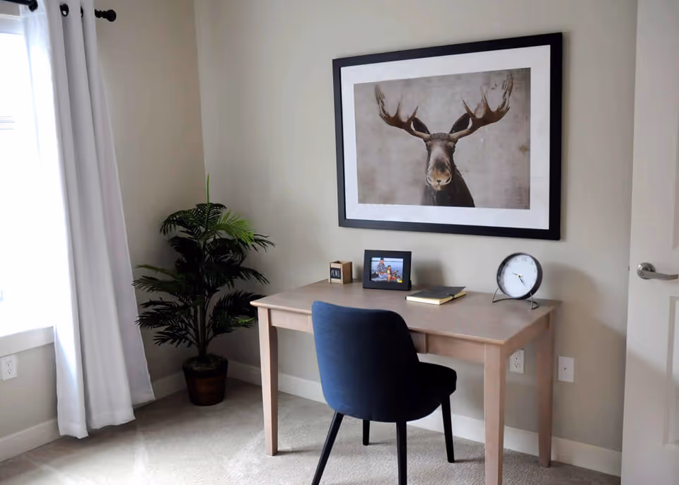 A small home office setup with a wooden desk and a dark blue chair. On the desk are a clock, a framed photo, a small block calendar, and a closed notebook. A large framed picture of a moose hangs on the wall above the desk. To the left, there is a window with white curtains and a potted plant in the corner.