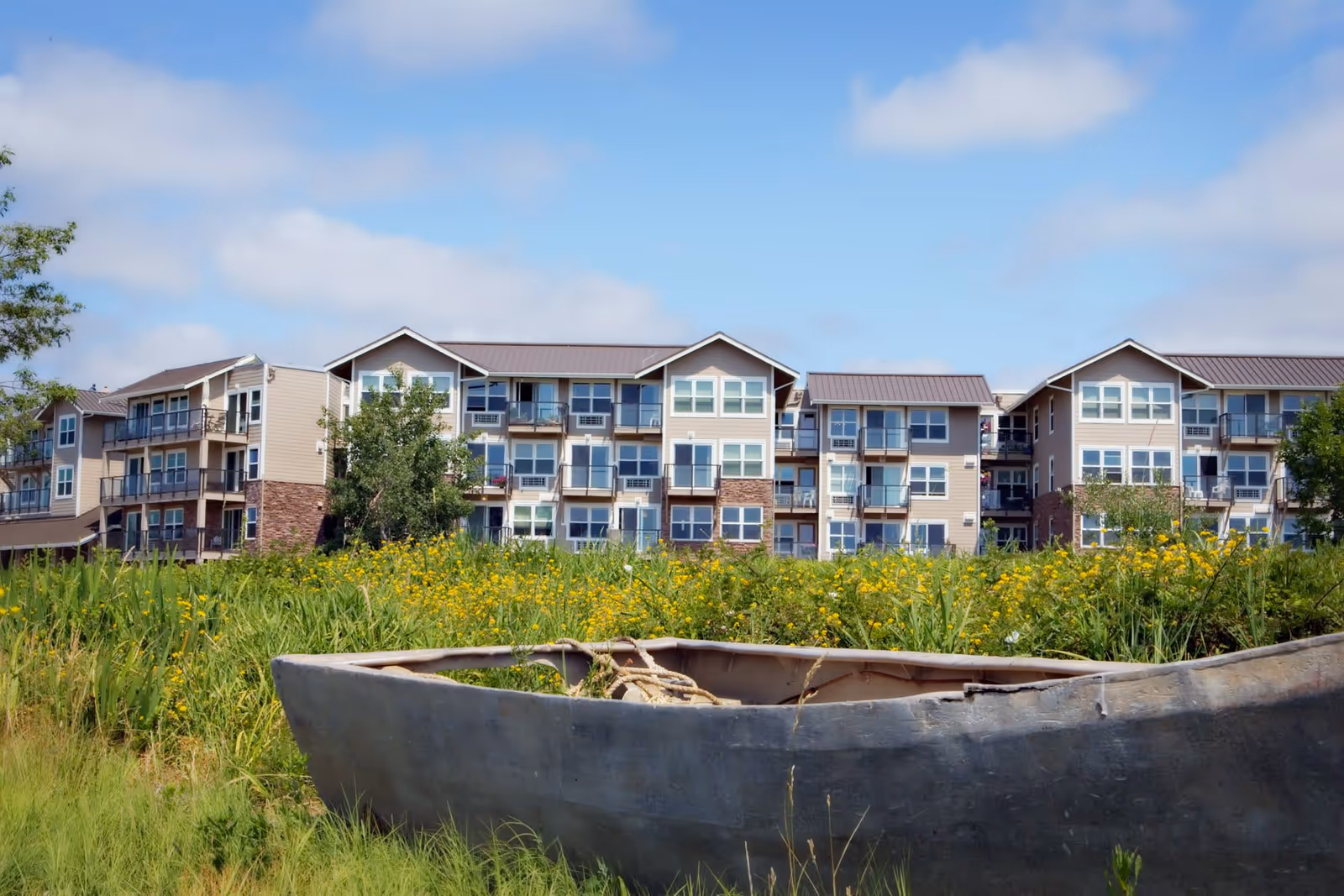 A multi-story residential building with balconies, surrounded by greenery and yellow wildflowers, under a partly cloudy blue sky. In the foreground, there is an old, weathered boat resting on the grass.