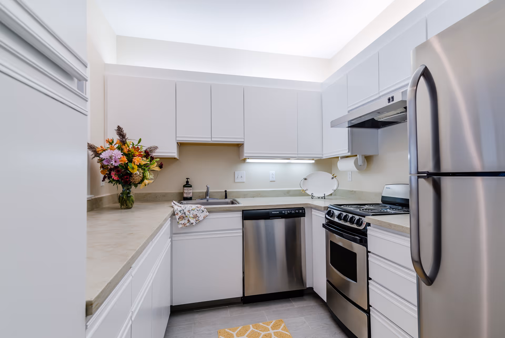 A modern kitchen with white cabinets, a stainless steel refrigerator, stove, and dishwasher. There is a vase with colorful flowers on the left countertop, a dish towel hanging near the sink, and a small decorative plate on a stand on the right countertop. The kitchen has under-cabinet lighting and a light-colored countertop.