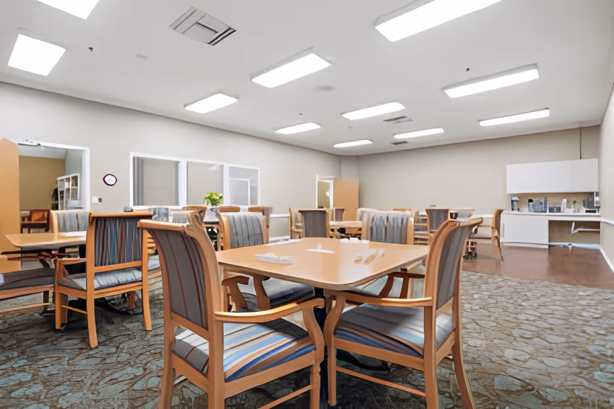 Bright communal dining room with wooden tables and striped upholstered chairs and a small kitchenette in the back.