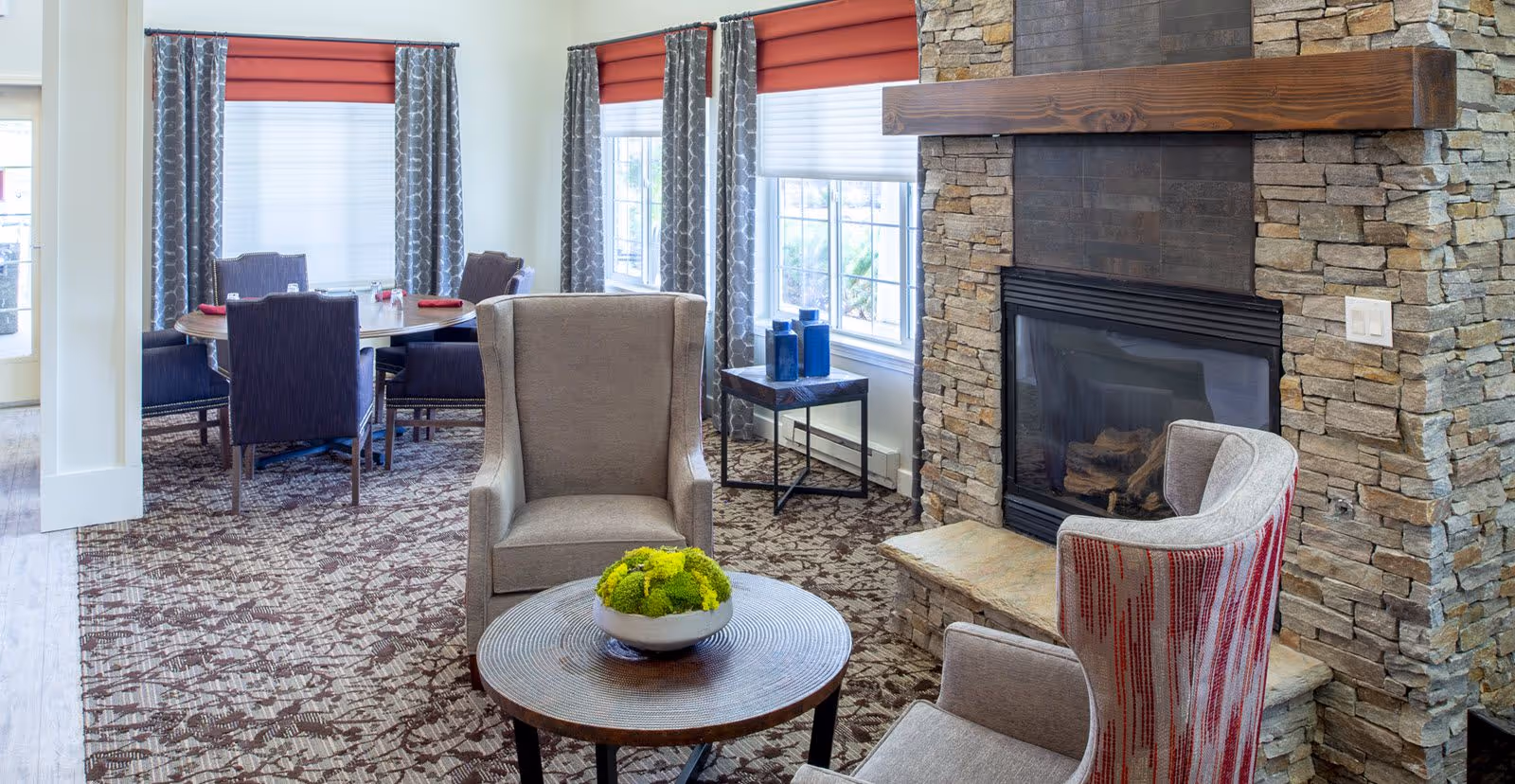 A cozy senior living common area with a stone fireplace, two upholstered armchairs, a round wooden coffee table with a green plant centerpiece, and a dining area with a round table and four chairs near windows with red and white blinds and patterned curtains.