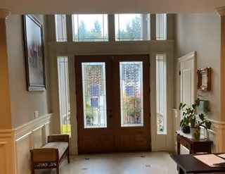 Interior view of an entryway with double wooden doors featuring decorative glass panels. The area is well-lit with natural light coming through the glass panels and sidelights. There is a bench on the left side, a small table with plants on the right, and framed artwork on the walls.