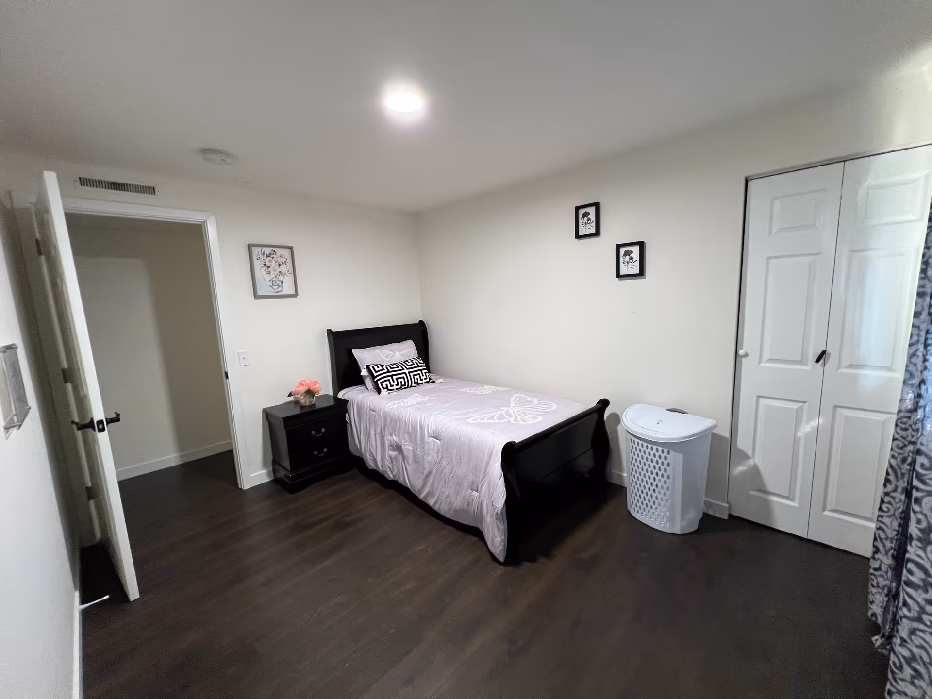 A small bedroom with dark wood flooring, a single black wooden bed with light purple bedding featuring a butterfly design, a black nightstand with a pink flower arrangement, a white laundry basket, white closet doors, and three framed floral pictures on the walls.