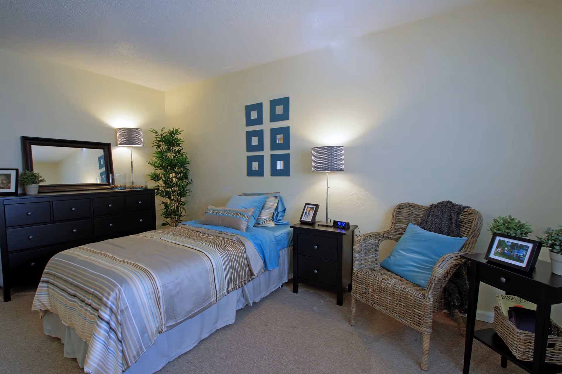 Neatly furnished bedroom with a single bed, dresser, nightstands, lamps, and a wicker chair with blue accents.