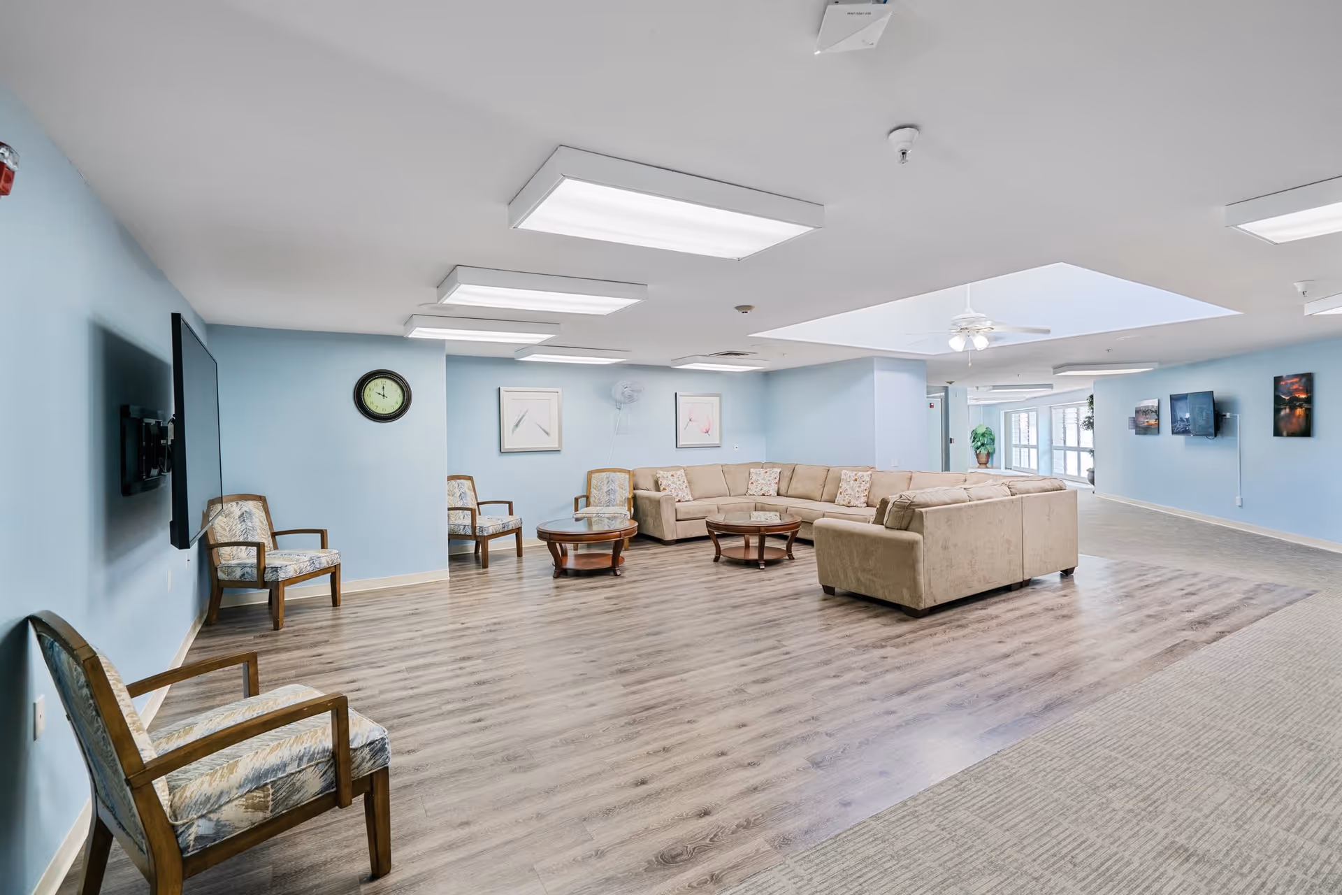 A spacious senior living facility common area with light blue walls and wood flooring. The room features a large beige sectional sofa, several wooden armchairs with patterned cushions, two round wooden coffee tables, a wall-mounted flat-screen TV, a clock, framed artwork, and ceiling lights. There is a skylight with a ceiling fan and a hallway leading to other areas.