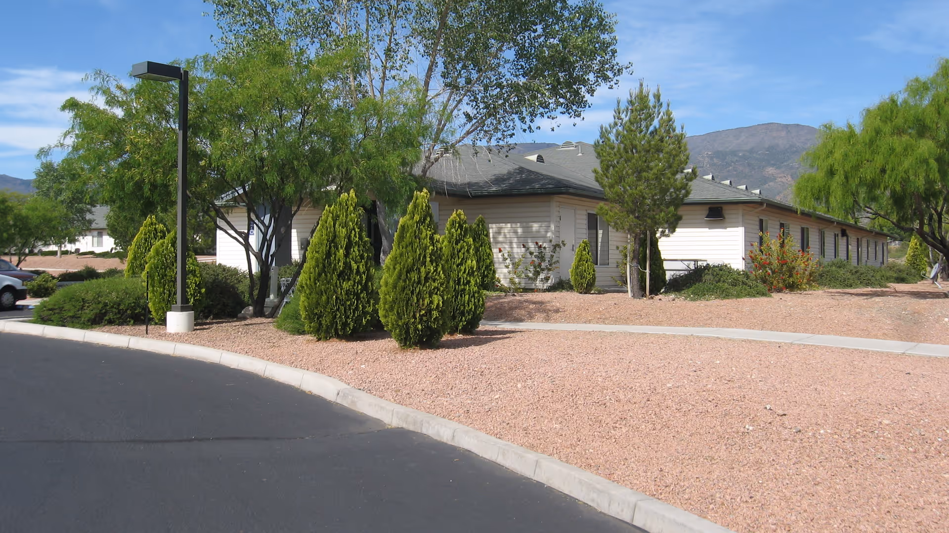 Single-story beige building with low desert landscaping, small trees and shrubs beside a curved driveway and distant mountains under a blue sky.