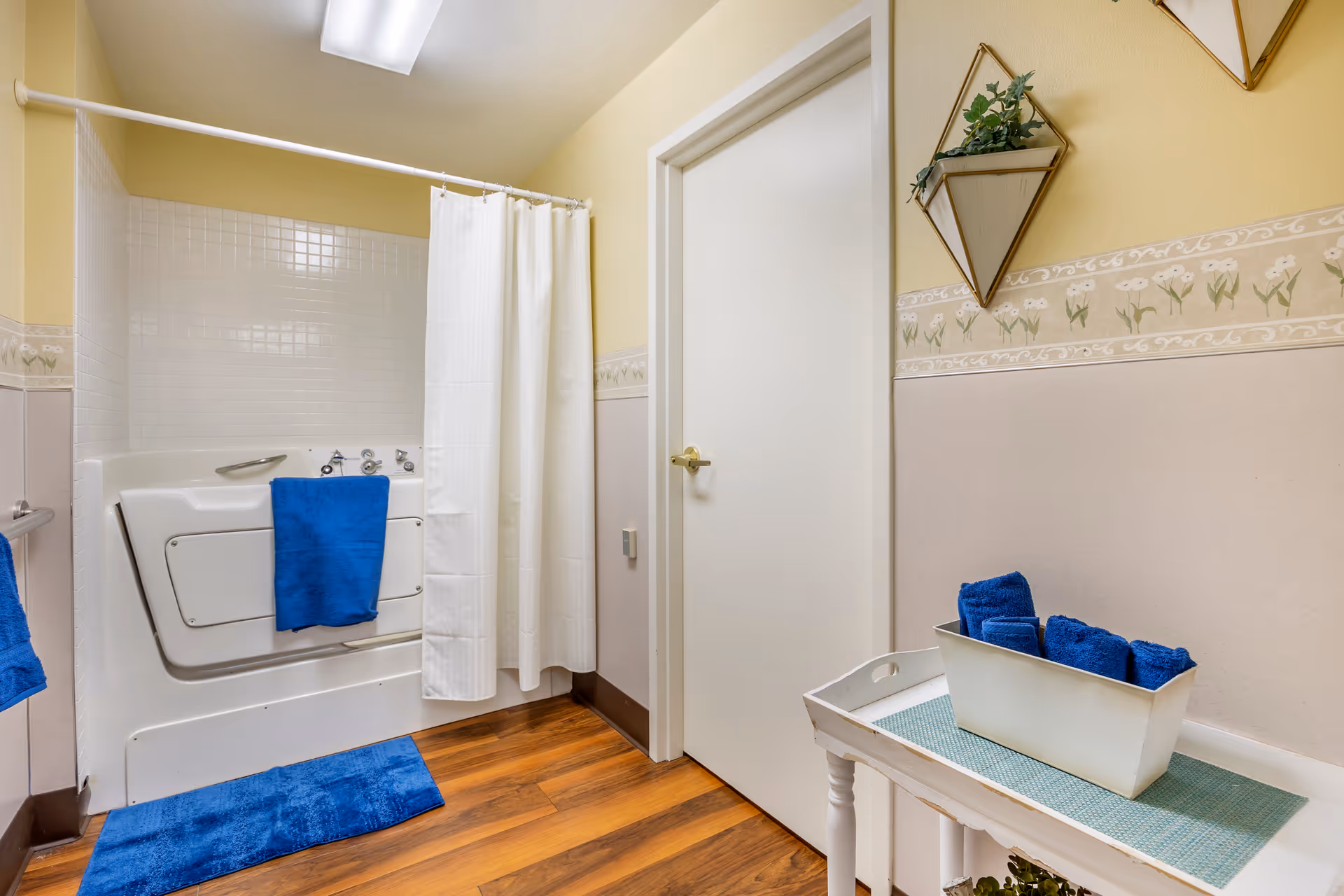 A bathroom with a walk-in bathtub featuring a white curtain and blue towels. The walls are painted yellow and beige with a floral border. There is a white door, a small white table with a container holding blue towels, and a wall-mounted plant holder with greenery.