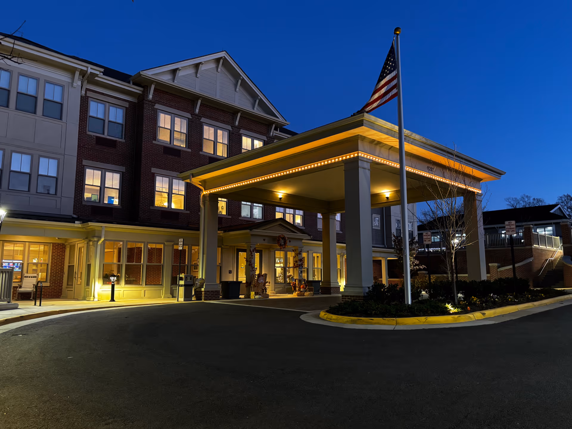 Exterior view of a multi-story senior living facility building at dusk with lights on inside. The entrance has a covered drop-off area with columns and an American flag on a flagpole. The sky is clear and dark blue.