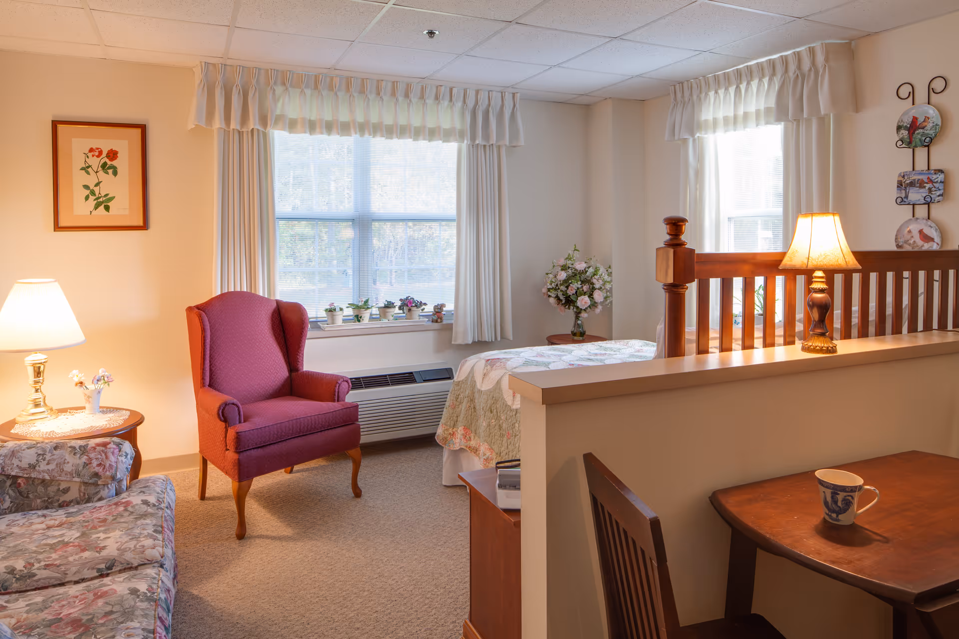 A cozy living area in a senior living facility with a red upholstered armchair, a floral patterned sofa, a wooden table with a cup, two table lamps, and a bed partially visible behind a half wall. The room has two windows with white curtains and several small potted plants on the windowsill. There is a framed flower artwork on the wall and a decorative wall hanging with three plates.