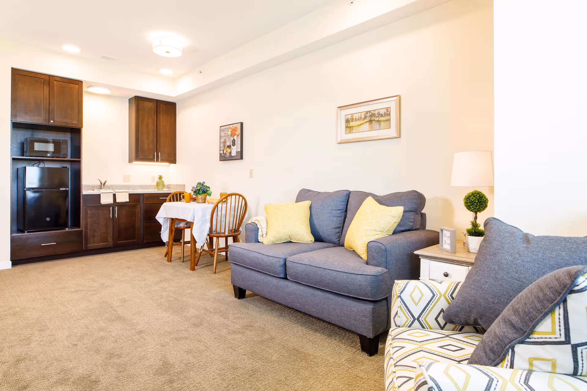 Bright living room with a gray loveseat, patterned armchair, small dining table, and a kitchenette with dark cabinets and a mini fridge.