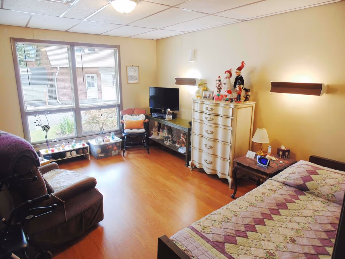 A cozy, well-lit bedroom with a single bed covered in a quilted blanket, a wooden nightstand with a lamp and alarm clock, a white dresser adorned with decorative figurines, a TV on a stand, a cushioned chair, and a large window letting in natural light.