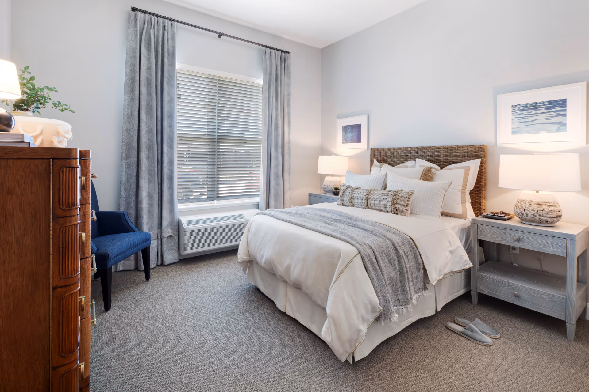 A cozy bedroom with a neatly made bed featuring white and beige bedding and multiple pillows. On either side of the bed are light gray wooden nightstands with matching lamps. A pair of gray slippers is placed on the carpeted floor next to the right nightstand. A wooden dresser and a blue upholstered chair are positioned near a window with gray curtains and white blinds. Two framed pictures hang on the light-colored walls above the nightstands.