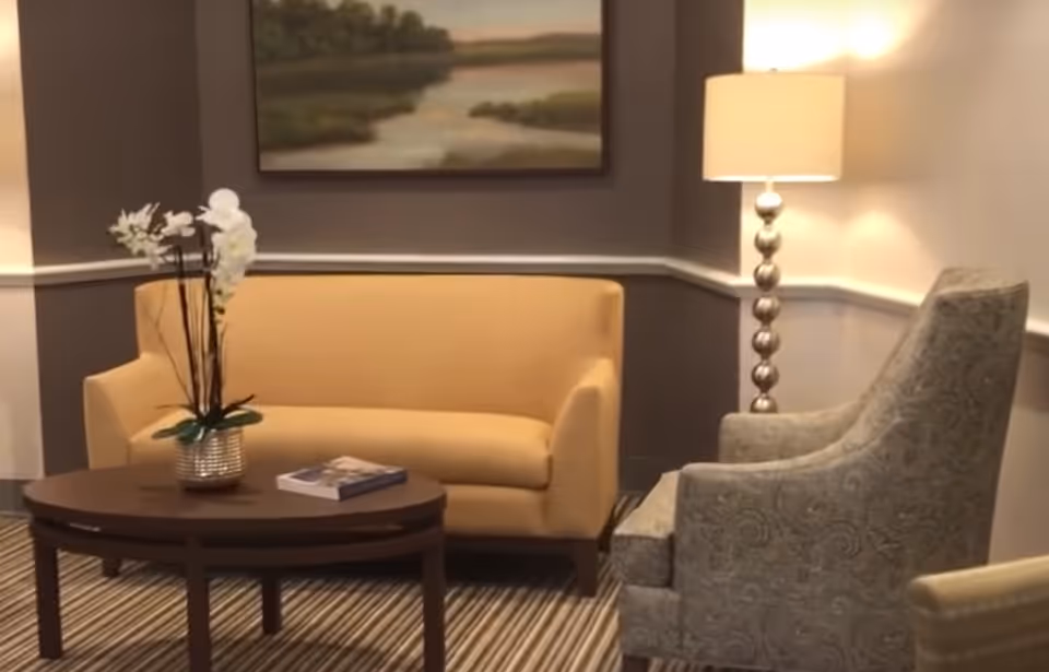 A seating area with a yellow sofa, patterned armchair, wooden coffee table holding a potted orchid and a book, a floor lamp, and wall art.