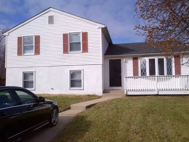 Exterior view of a two-story white house with red shutters and a small front porch with a white railing. A black car is parked on the driveway leading up to the house. There is a tree with brown leaves on the right side and a grassy lawn in front.