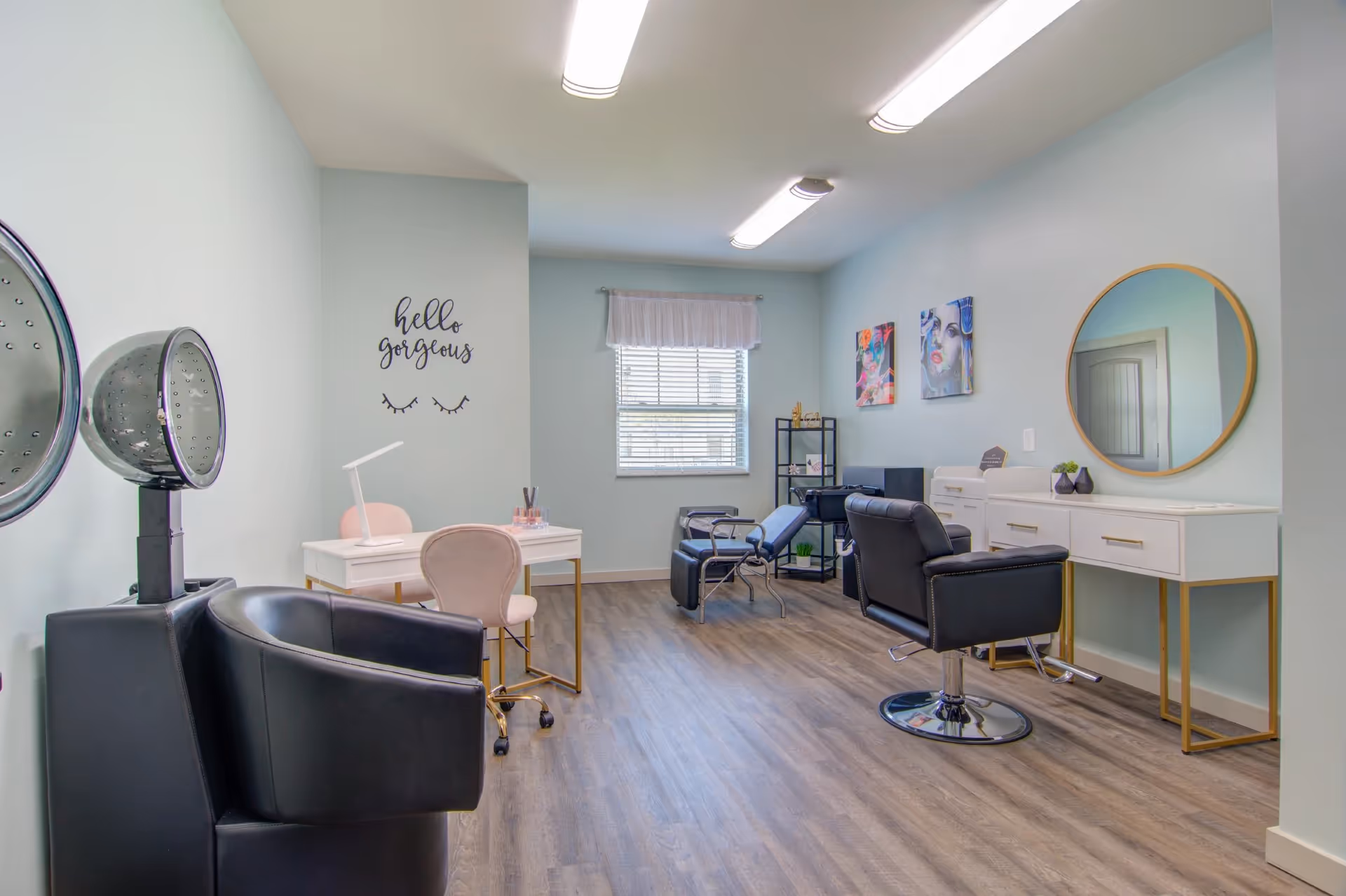 A bright salon-style room with styling chairs, hooded hair dryers, manicure stations, a round mirror and a wall decal reading 'hello gorgeous'.