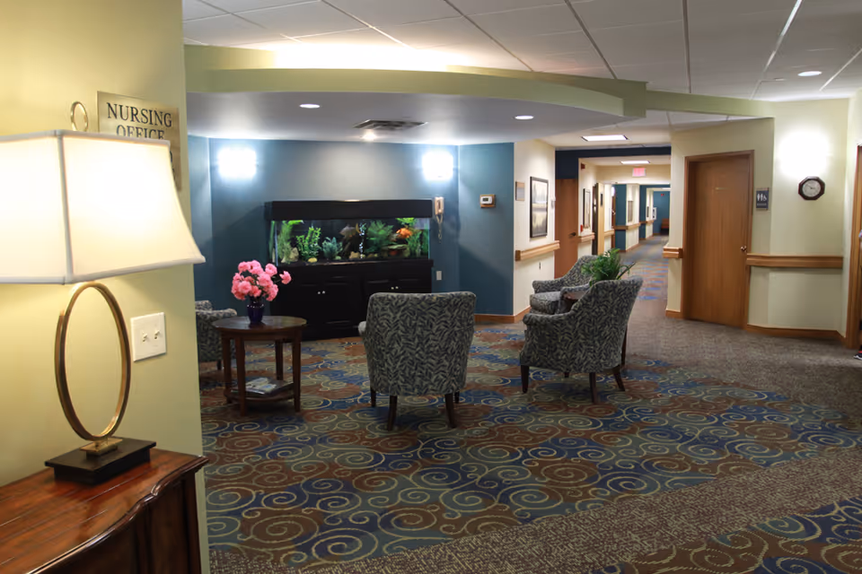 Interior view of a senior living facility hallway with a seating area featuring four patterned armchairs around a small round table with a vase of pink flowers. There is a large fish tank against a blue accent wall, a lamp on a wooden table in the foreground, and a sign indicating the nursing office. The hallway extends into the background with doors and framed pictures on the walls.