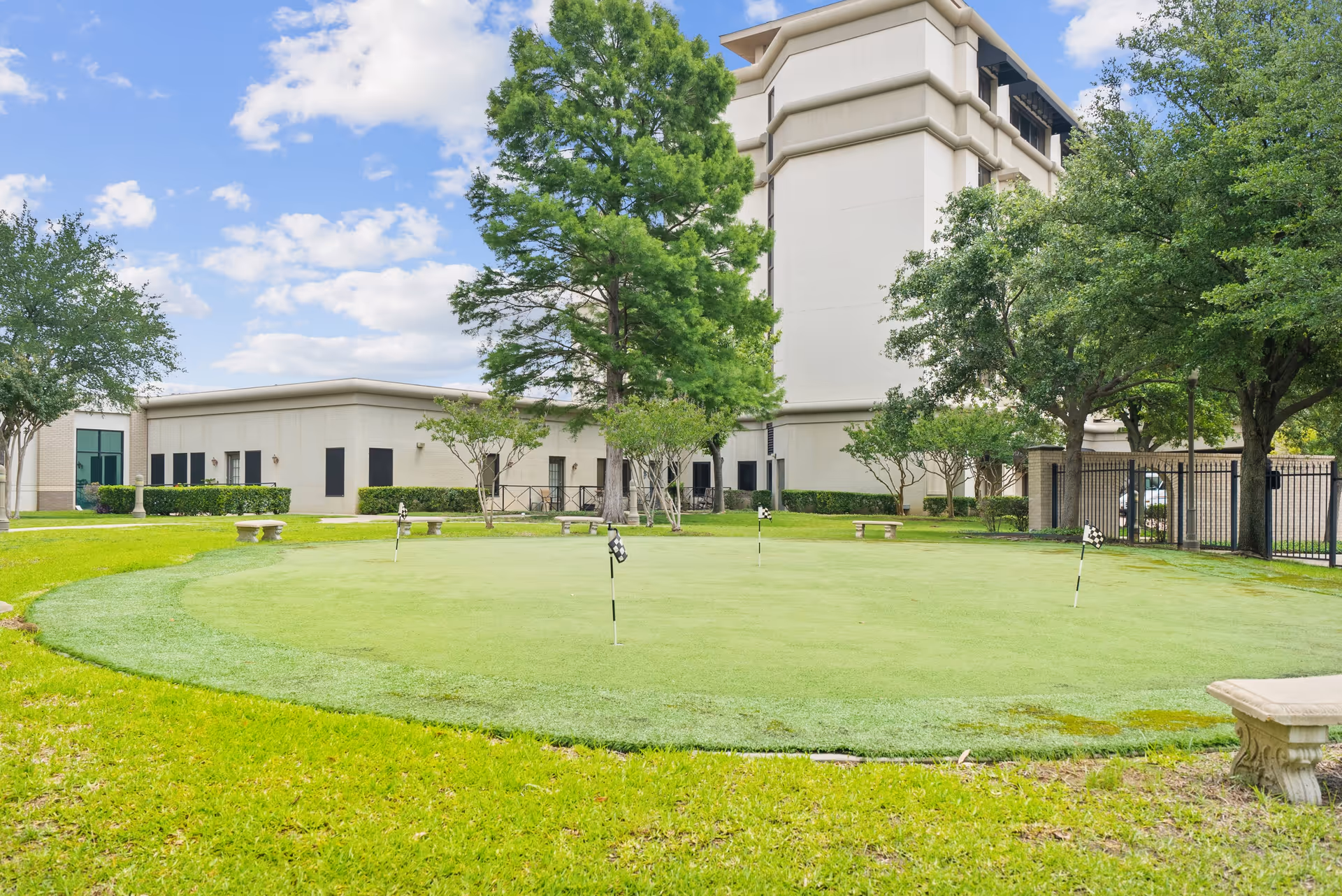 Outdoor putting green surrounded by grass and trees with benches around it, adjacent to a multi-story building under a partly cloudy sky.