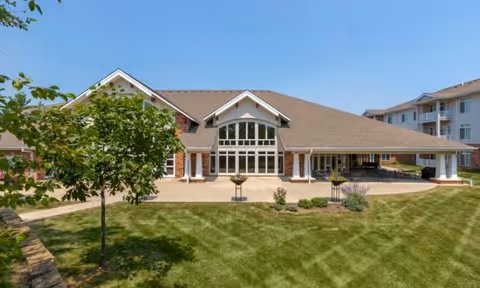 Exterior view of a senior living facility building with a large covered patio area, multiple windows, and a well-maintained lawn with a small tree and landscaping in front.