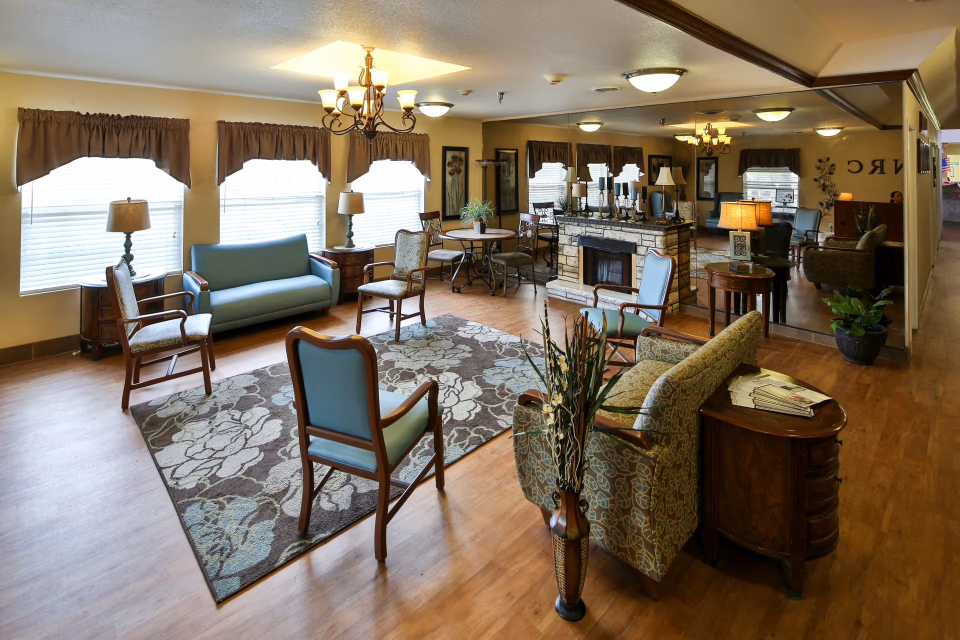 Bright communal lounge in a senior living facility with sofas, chairs, a rug, lamps, and a stone fireplace.
