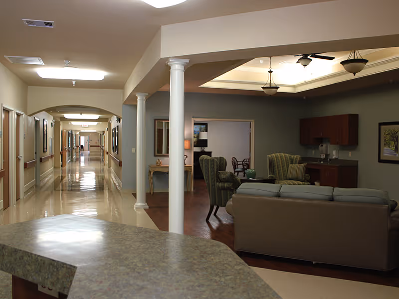 Interior view of a senior living facility hallway with a seating area to the right. The hallway has multiple doors along the left side and a shiny floor. The seating area includes a sofa, armchairs, a small table, and a kitchenette with cabinets. The ceiling has recessed lighting and hanging light fixtures.