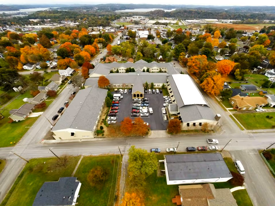 Aerial view of River Oaks Place - Lenoir City facility surrounded by residential houses and trees with autumn foliage. The facility consists of multiple connected buildings with a parking lot filled with cars in the center. Roads and green lawns are visible around the property.