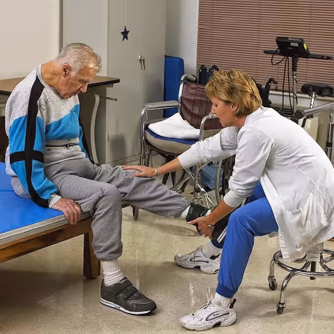 A healthcare professional assists an elderly man seated on a bench by examining or stretching his leg in a clinical or rehabilitation room equipped with medical and mobility equipment.