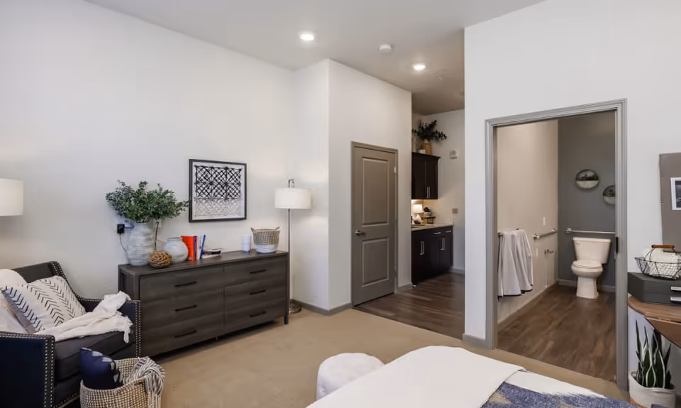 A cozy bedroom area with a dark wooden dresser adorned with decorative items and a framed artwork above it. To the left, there is a comfortable armchair with pillows and a throw blanket. The room has beige carpet flooring and white walls. To the right, there is an open doorway leading to a bathroom with a toilet, grab bars, and wood-style flooring. Another doorway leads to a small kitchenette area with dark cabinets and a countertop.