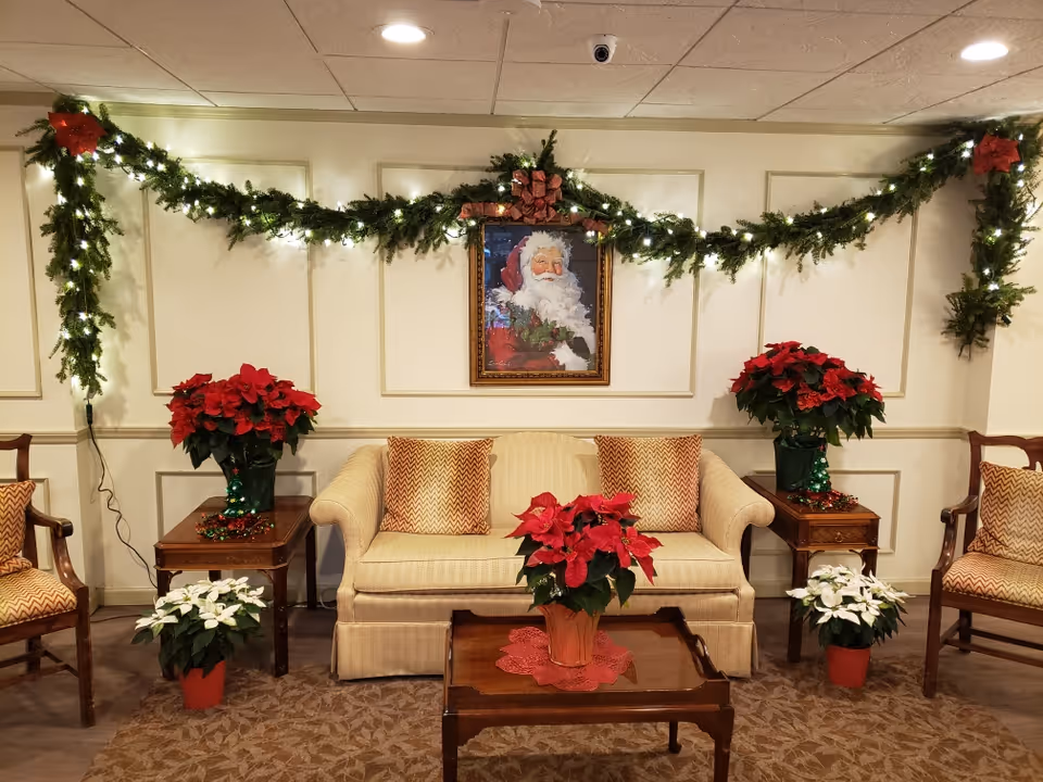 A cozy living room decorated for Christmas with a beige sofa flanked by two wooden side tables holding red poinsettia plants. Two matching armchairs are placed on either side. A wooden coffee table in front of the sofa holds another red poinsettia. Garland with lights and red bows is draped across the wall above the sofa, which features a framed picture of Santa Claus. The floor is carpeted with a patterned design.