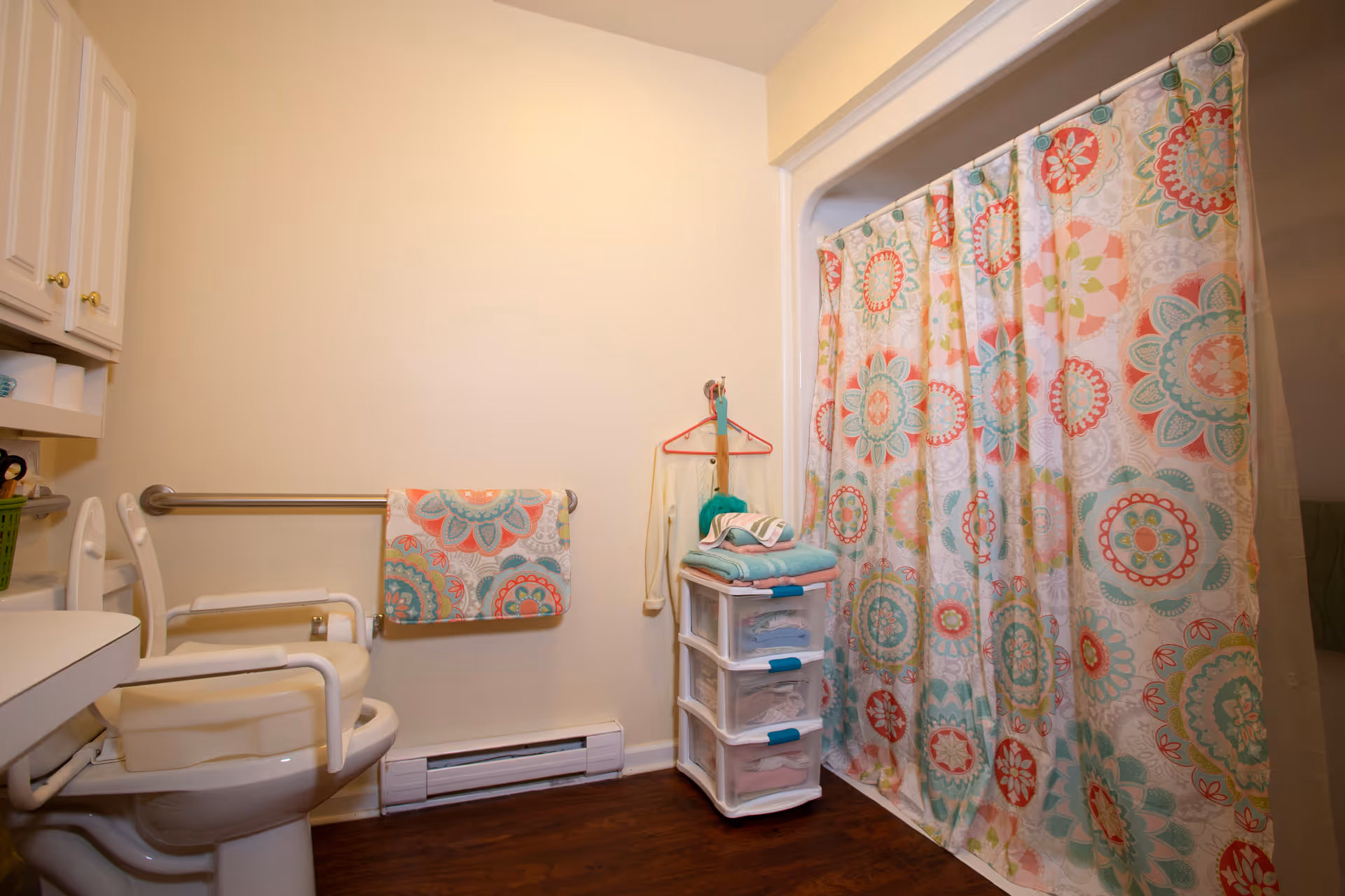 A bathroom with a white toilet equipped with safety rails, a colorful floral shower curtain, a towel rack with a matching floral towel, a small plastic drawer unit holding folded towels and clothes, and a wooden floor.