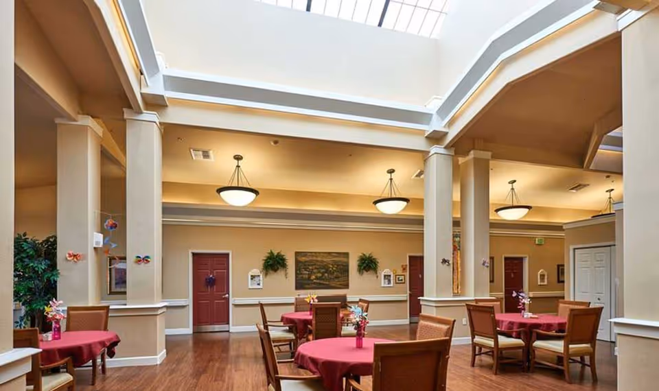 Spacious assisted living dining area with round tables covered in red tablecloths, wooden chairs, tall pillars, a large skylight, and warm pendant lighting.