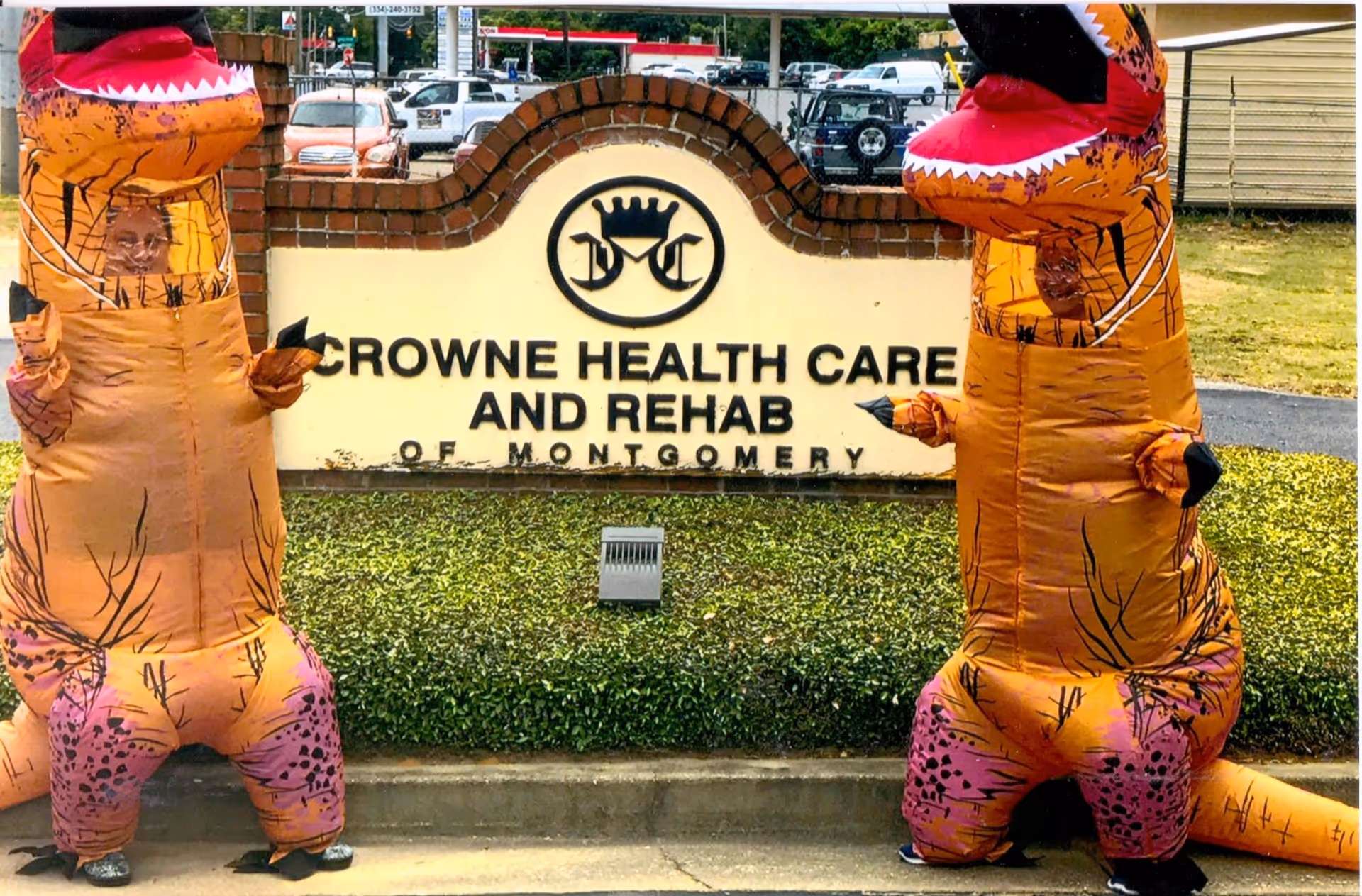 Two people dressed in inflatable dinosaur costumes standing on either side of a brick and stone sign that reads 'Crowne Health Care and Rehab of Montgomery' with a logo above the text. The background shows parked cars and some greenery.