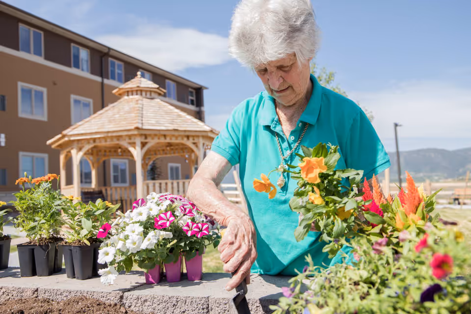 An elderly woman wearing a teal shirt is gardening outdoors, planting colorful flowers in a raised garden bed. Behind her is a wooden gazebo and a multi-story building under a clear blue sky.