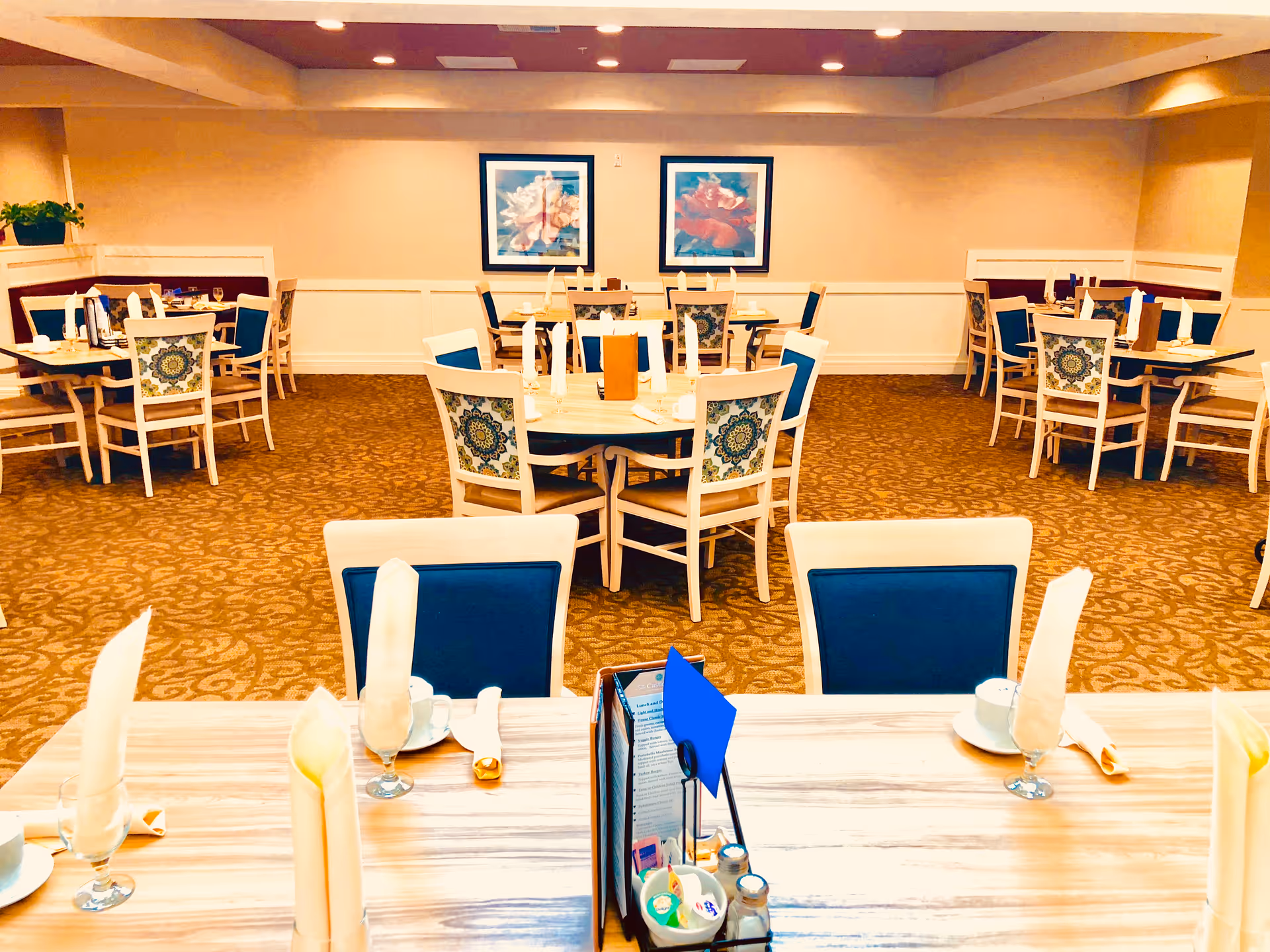 A dining room in a senior living facility with multiple tables and chairs arranged neatly. Each table is set with folded napkins, cups, and glasses. The chairs have blue cushions and patterned backs. The room has beige walls with two framed floral paintings and a patterned carpet. The lighting is warm and ceiling lights are visible.