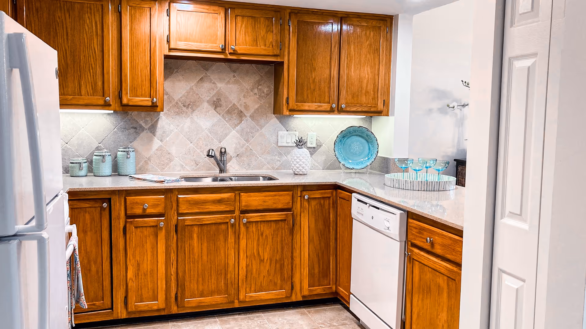 A kitchen with wooden cabinets, a white refrigerator, a white dishwasher, a double sink, and a tiled backsplash. The countertop has decorative items including a white pineapple, a blue decorative plate, a tray with blue glasses, and three small jars. The floor is tiled and the walls are painted white.