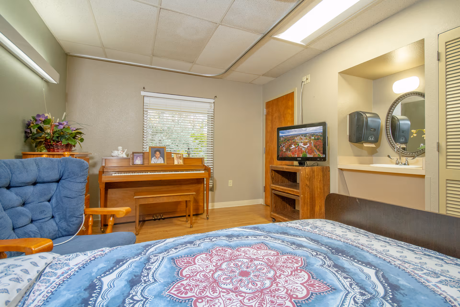 A cozy room with a bed covered in a blue and red patterned blanket, a blue cushioned chair with wooden armrests, a wooden piano with framed photos on top, a small TV on a wooden stand, and a sink area with a round mirror and soap dispenser.